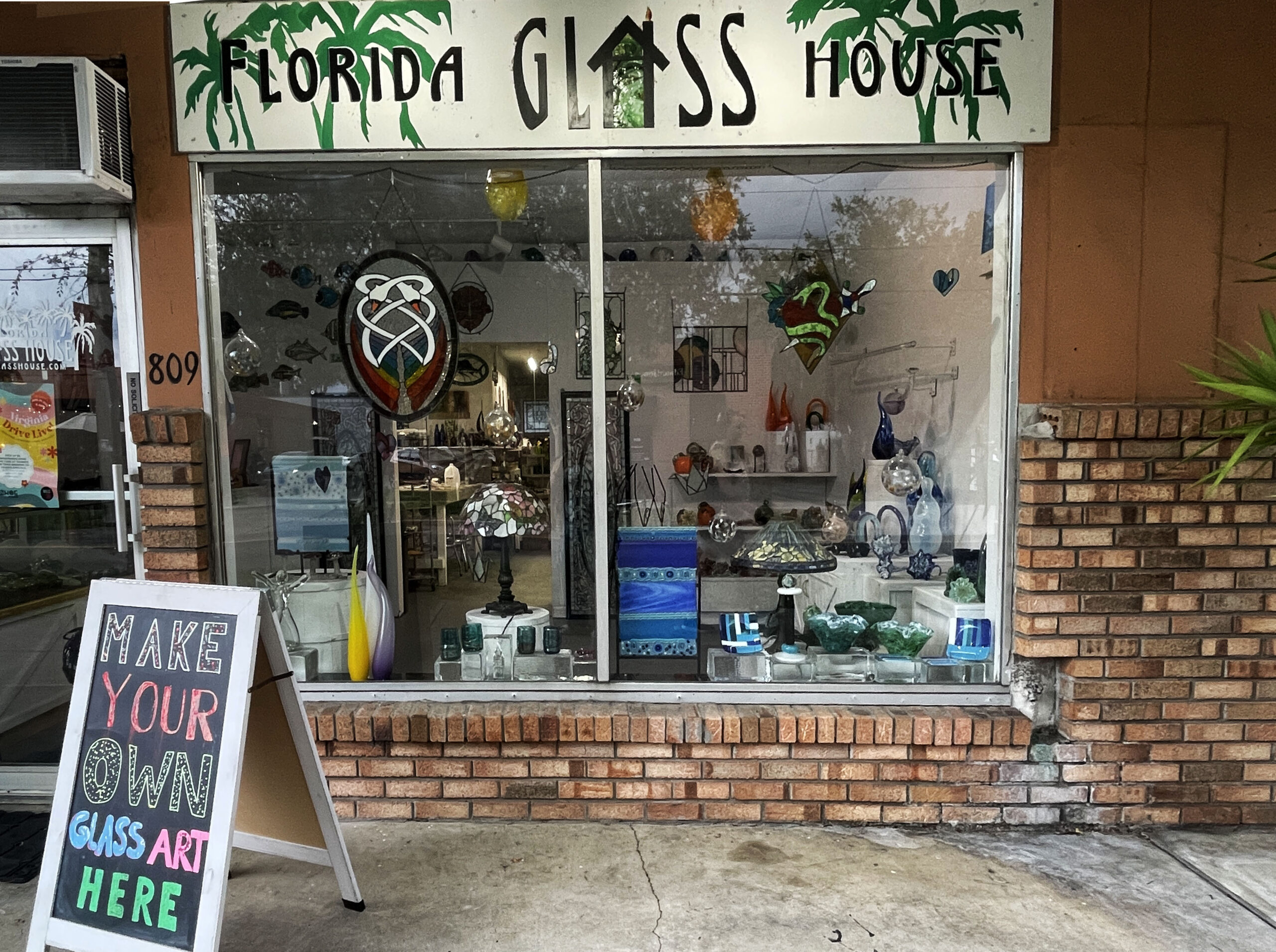 Florida Glass House storefront. Brick wall with a large display window with glass art. Signs read 'Florida Glass House,' and 'Make your own glass art here.'
